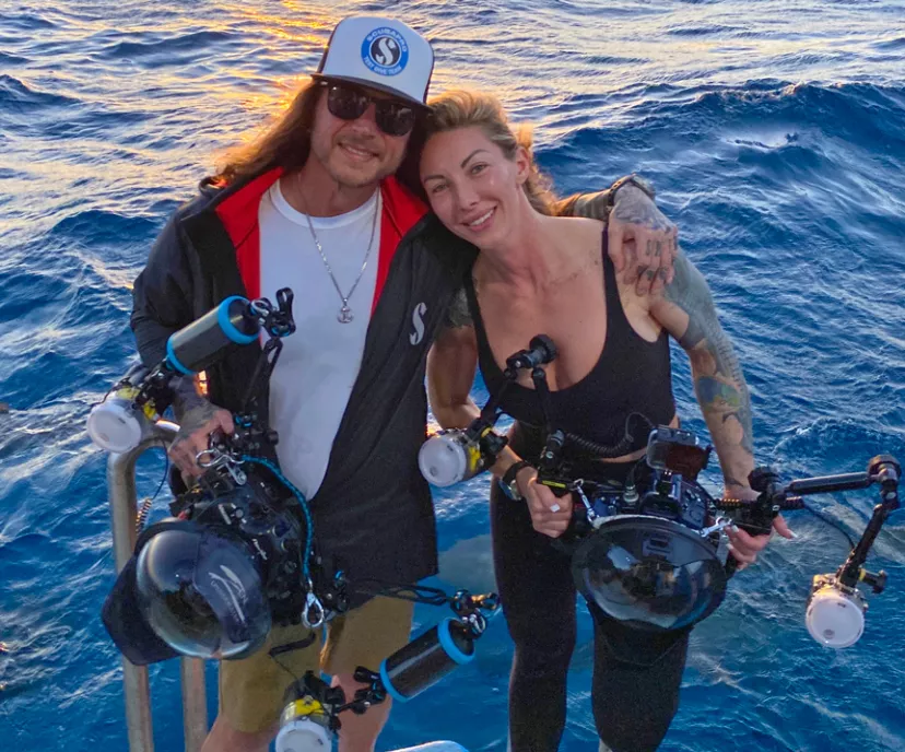 Deano and Erin Cook holding large underwater camera rigs with the ocean behind them.